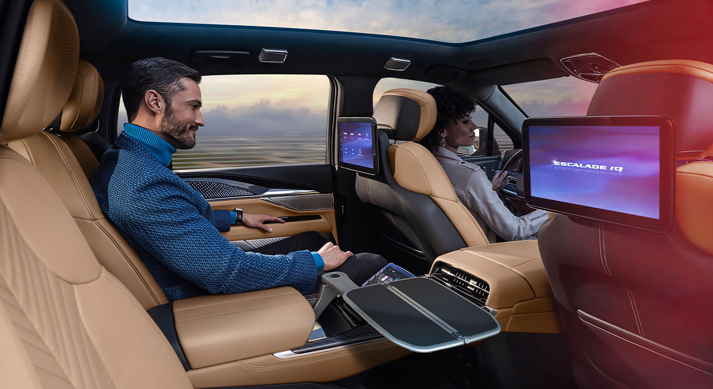 Luxury SUV interior showing two people seated in tan leather seats. They face forward, with entertainment screens displaying “Escalade IQ.” The mood is relaxed.